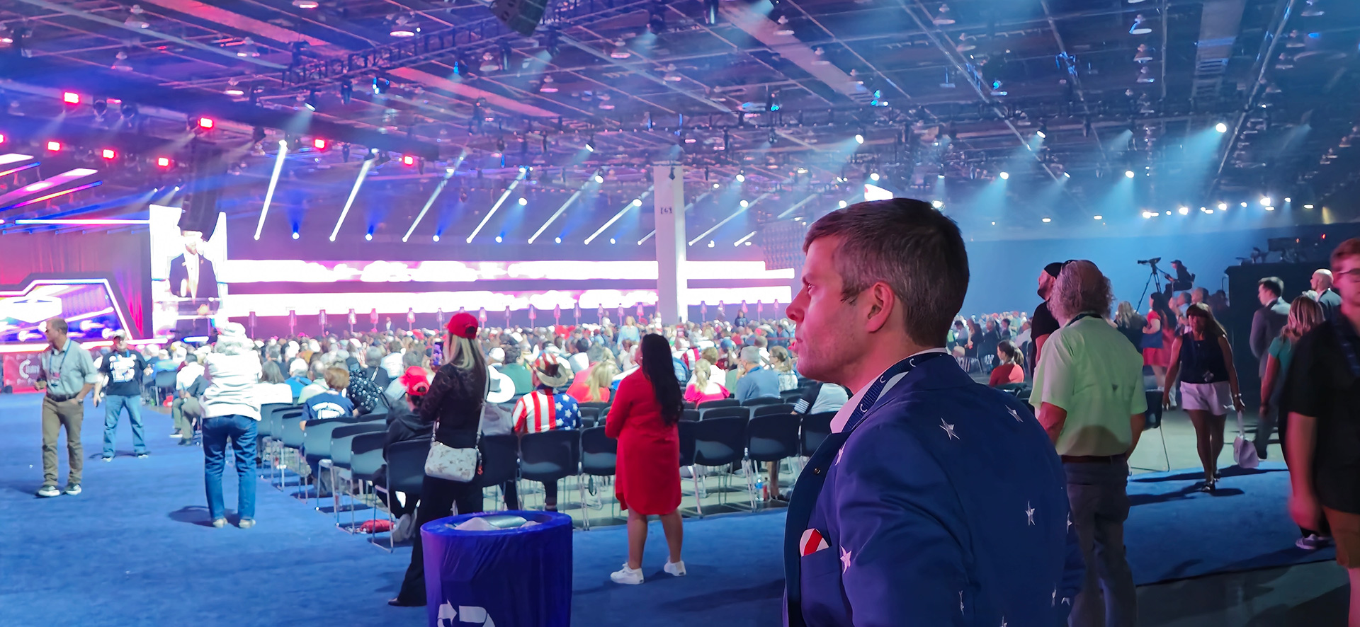 Stephen Schumacher in profile, looking toward the stage in a large event hall.